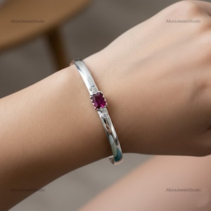 Könnte beinhalten: Ein silbernes Armreif-Armband mit einem rechteckigen, tiefroten Edelstein in der Mitte, flankiert von zwei kleineren, runden, klaren Edelsteinen. Das Armband wird am Handgelenk getragen.