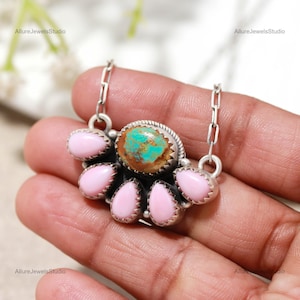 May include: A silver necklace featuring a central oval turquoise stone with brown matrix, surrounded by six teardrop-shaped pink stones in serrated bezels. The pendant is attached to a silver chain with rectangular links, creating a fan-like arrangement.