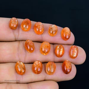 Op de afbeelding: Een verzameling van 12 ovale zonnesteen cabochons, vari&euml;rend in kleur van oranje tot rood-oranje. De stenen zijn glad en gepolijst, met een lichte schittering.