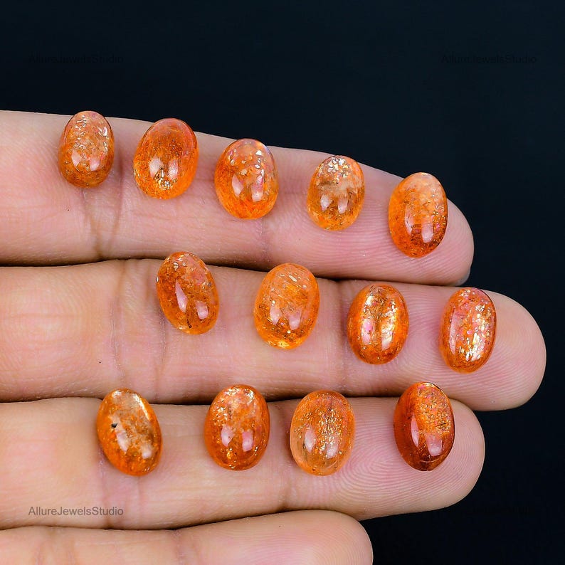 Op de afbeelding: Een verzameling van 12 ovale zonnesteen cabochons, vari&euml;rend in kleur van oranje tot rood-oranje. De stenen zijn glad en gepolijst, met een lichte schittering.