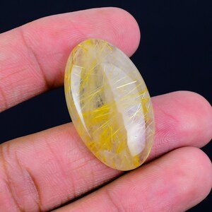 May include: A clear oval-shaped gemstone with gold rutilated inclusions. The gemstone is set in a cabochon cut and has a smooth, polished surface.