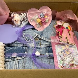 May include: A denim jacket, a purple heart-shaped item, and various accessories are arranged inside a cardboard box. The accessories include a bunny figurine, a hair clip, and a pink bag with sweets. The box is lined with pink tissue paper.