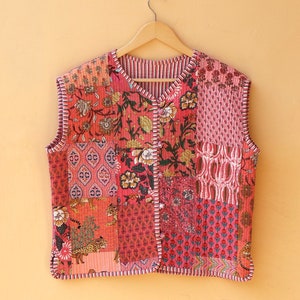 Peut inclure: Un gilet matelassé sans manches avec un motif patchwork dans des tons de rose, rouge et orange. Le gilet a un motif floral et est suspendu à un cintre en bois.