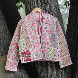 Puede incluir: Chaqueta acolchada con diseño patchwork. Presenta varios estampados florales en tonos rosa, verde y rojo, sobre un fondo neutro. La chaqueta está colgada en una percha de madera frente a un tronco de árbol.