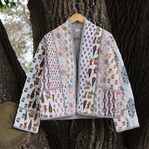 May include: A patchwork quilted jacket with floral and geometric patterns in pink, blue, and green. The jacket has a striped lining and is suspended on a wooden hanger. The jacket's design is a mix of patterns.