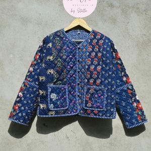 May include: A quilted jacket in shades of blue, featuring a patchwork design with floral and animal motifs. The jacket has two front pockets and a striped lining. The brand name "sandria by Stella" is visible on a round tag.