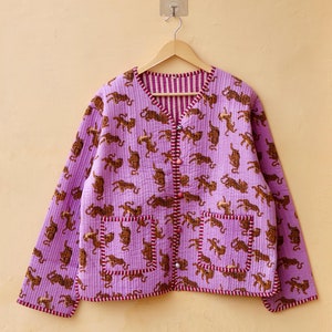 Könnte beinhalten: Eine lavendelfarbene Steppjacke mit braunem Tigerdruck. Die Jacke hat einen V-Ausschnitt, zwei Vordertaschen und braune Knöpfe. Die Paspel und das Innenfutter sind gestreift. Die Jacke hängt an einem Holzbügel.