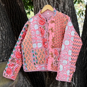 May include: A quilted jacket with a patchwork design in shades of pink, peach, and red. The jacket features floral and animal motifs, with a striped lining and a button-up front. The jacket is hanging on a wooden hanger.
