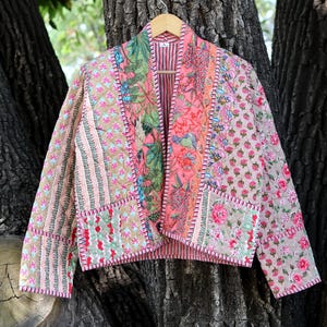 May include: A patchwork jacket with a floral and striped pattern in shades of pink, green, and beige. The jacket is hanging on a wooden hanger against a tree trunk background. The jacket has a quilted design.