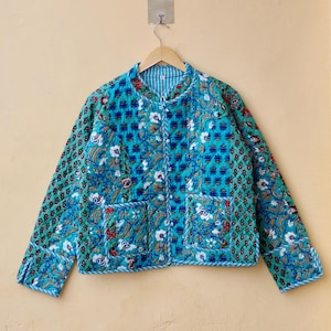 Könnte beinhalten: Eine tealblaue gesteppte Jacke mit Blumendruck und weißem Besatz. Die Jacke hat einen Stehkragen und einen Knopfverschluss. Sie hat zwei aufgesetzte Taschen auf der Vorderseite.