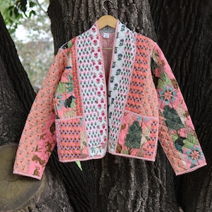 Puede incluir: Chaqueta acolchada con diseño patchwork. La chaqueta presenta varios patrones, incluyendo motivos florales y geométricos, en tonos rosa, verde y blanco. Tiene cuello, bolsillos y está colgada de una percha de madera.