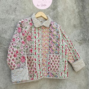 Pode incluir: Casaco acolchoado com padrões florais e listrados. O casaco apresenta uma mistura de estampas florais rosa, verde e branco, com punhos e gola listrados. O casaco tem dois bolsos e está pendurado em um cabide de madeira.