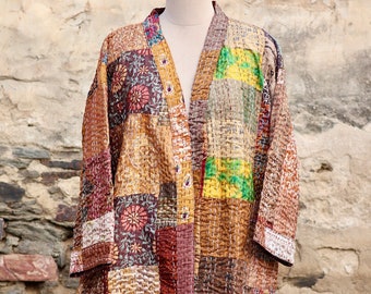 May include: A patchwork kimono-style jacket in warm tones of brown, gold, and burgundy. The jacket features a variety of patterned fabric squares, stitched together with visible white thread. It is displayed on a mannequin against a textured stone wall.