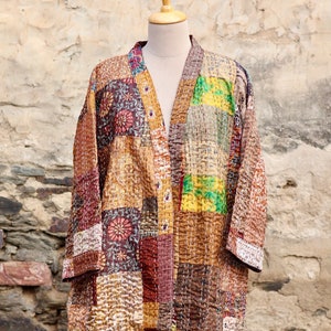 Puede incluir: Una chaqueta estilo kimono patchwork en tonos cálidos de marrón, dorado y burdeos. La chaqueta presenta una variedad de cuadrados de tela estampada, cosidos con hilo blanco visible. Se exhibe en un maniquí contra una pared de piedra texturizada.