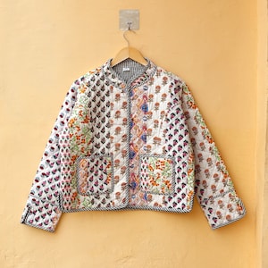 May include: A white quilted jacket with a patchwork design featuring floral patterns in various colors, including pink, orange, and blue. The jacket has a zipper closure and two pockets.