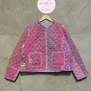 Könnte beinhalten: Gesteppte Jacke in Rosa mit floralen und geometrischen Mustern. Sie hat einen runden Kragen, zwei Vordertaschen und einen Knopfverschluss. Die Jacke hängt an einem Holzbügel. Der Text "sandra boutique by Stella" befindet sich in einem Kreis darüber.