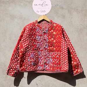 Op de afbeelding: Rood gewatteerd jack met een bloemenprint en een knoopsluiting. Het jack hangt aan een houten hanger. Het label op de hanger luidt "sandra boutique in by Stella".