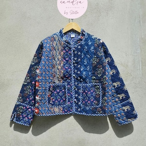 Könnte beinhalten: Eine gesteppte blaue Jacke mit einem Patchwork-Design mit floralen und tierischen Motiven. Die Jacke hat einen Stehkragen, Knopfverschlüsse und zwei Vordertaschen. Die Bordüre ist aus weiß-blauem Seil. Die Jacke hängt an einem Holzbügel.