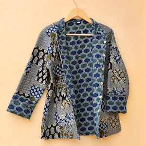 Könnte beinhalten: Eine gesteppte Jacke mit blauem und grünem Muster und Knopfverschluss. Die Jacke hängt an einem Holzbügel an einer hellorangen Wand.