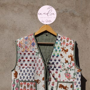 Könnte beinhalten: Gesteppte Weste mit farbenfrohem Blumen- und Tigermuster. Die Weste hat einen V-Ausschnitt, zwei Taschen und ein grün gestreiftes Futter. Sie hängt an einem Holzbügel. Der Markenname "sandria boutique in by Stella" ist sichtbar.