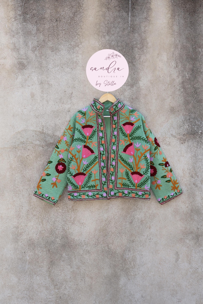 May include: A green embroidered jacket with pink, orange, and purple floral designs. The jacket has a mandarin collar and a front zipper closure. The jacket is hanging on a hanger against a textured wall. The text "sandra boutique in by Stella" is on a pink circle behind the jacket.