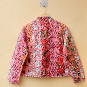 Op de afbeelding: Een gewatteerde jas met een patchwork ontwerp met bloemen- en paisley patronen in roze, rode en oranje tinten. De jas hangt aan een houten hanger tegen een beige muur.