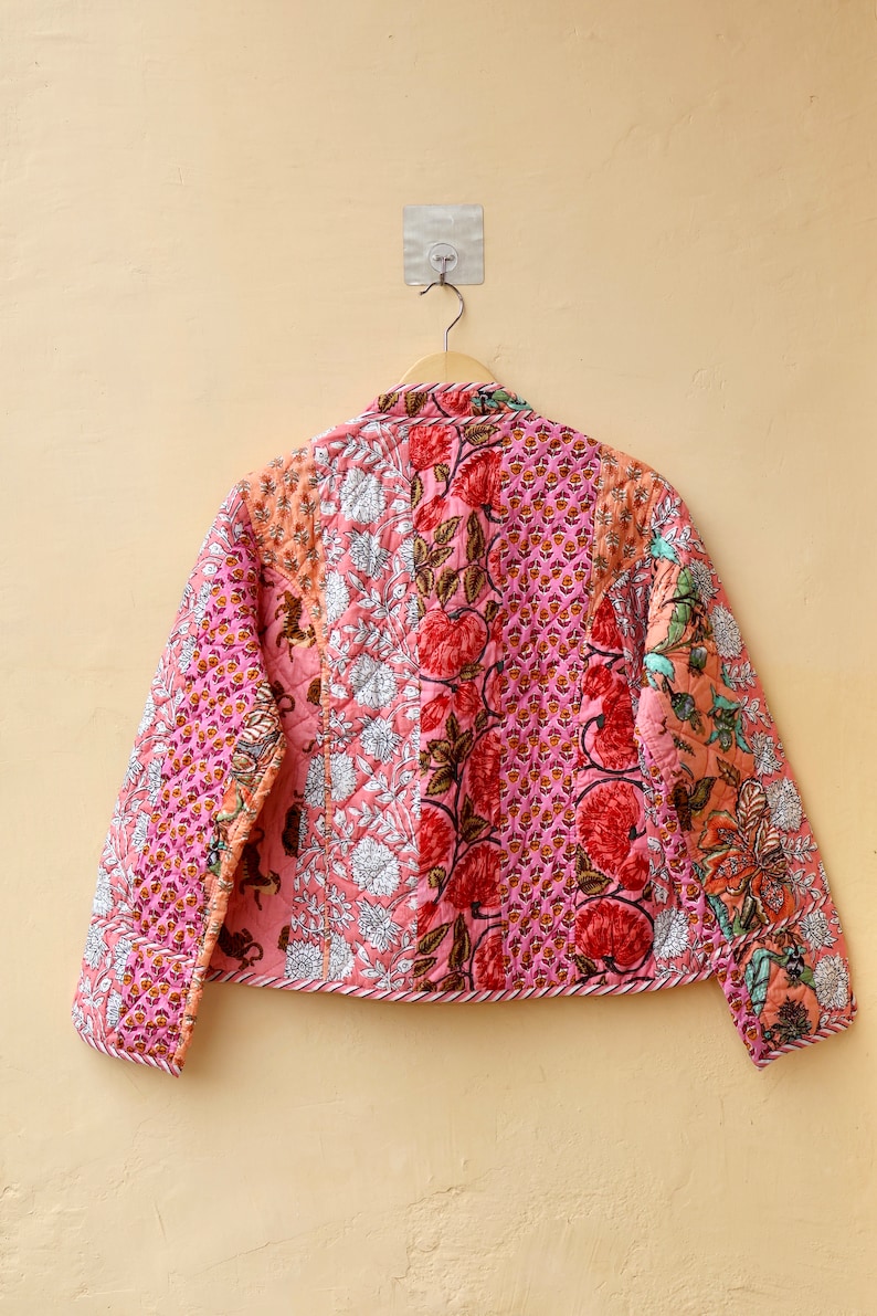 Op de afbeelding: Een gewatteerde jas met een patchwork ontwerp met bloemen- en paisley patronen in roze, rode en oranje tinten. De jas hangt aan een houten hanger tegen een beige muur.