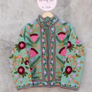 May include: A mint green embroidered jacket with a floral pattern in shades of pink, orange, and green. The jacket has a mandarin collar and a front closure. The label on the hanger reads "sandra boutique in by Stella."