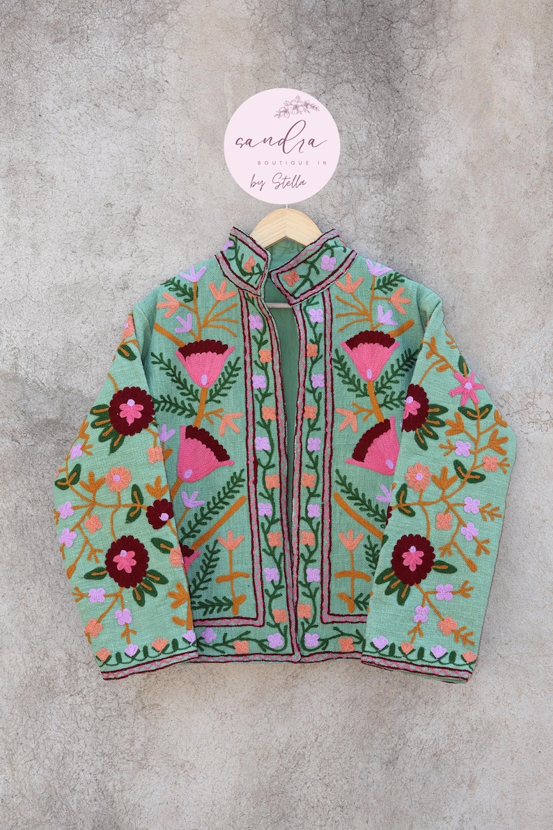 May include: A mint green embroidered jacket with a floral pattern in shades of pink, orange, and green. The jacket has a mandarin collar and a front closure. The label on the hanger reads "sandra boutique in by Stella."