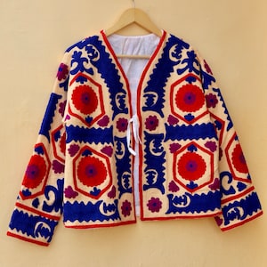 May include: A cream-colored jacket with a blue, red, and purple floral embroidery pattern. The jacket has a V-neck and a tie closure at the front.