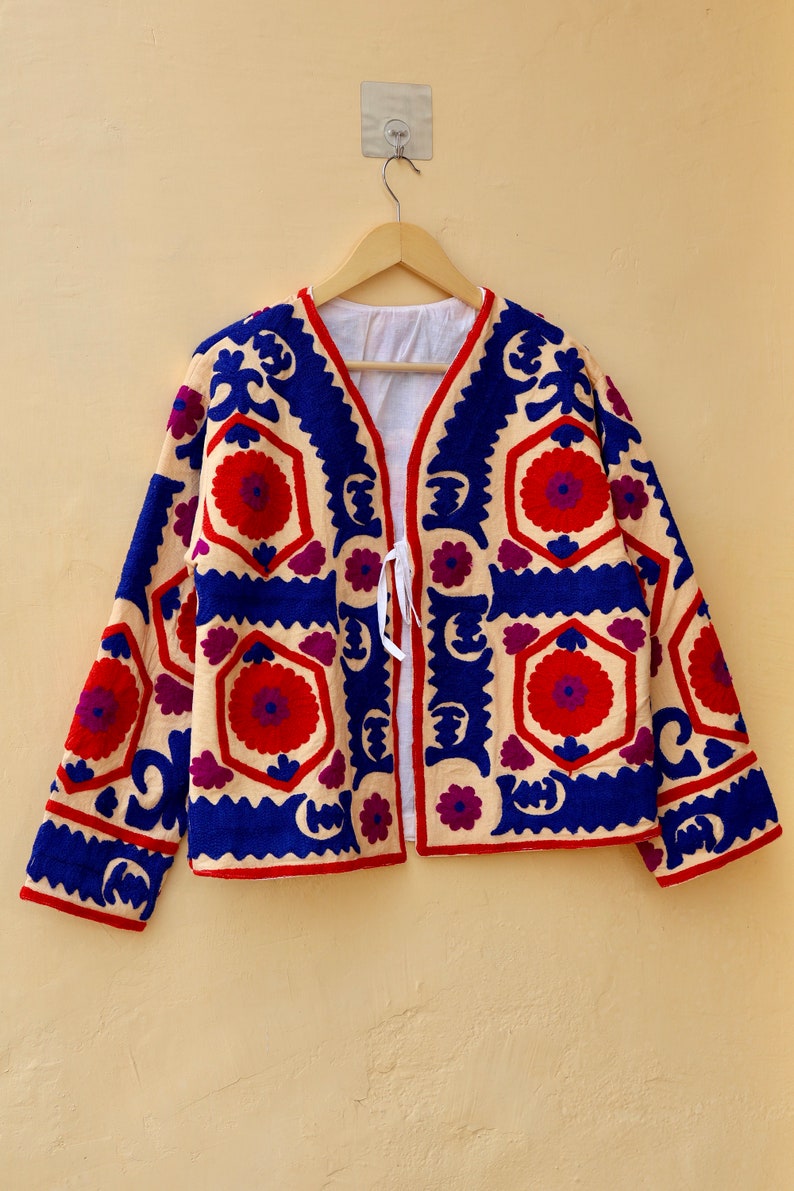 May include: A cream-colored jacket with a blue, red, and purple floral embroidery pattern. The jacket has a V-neck and a tie closure at the front.