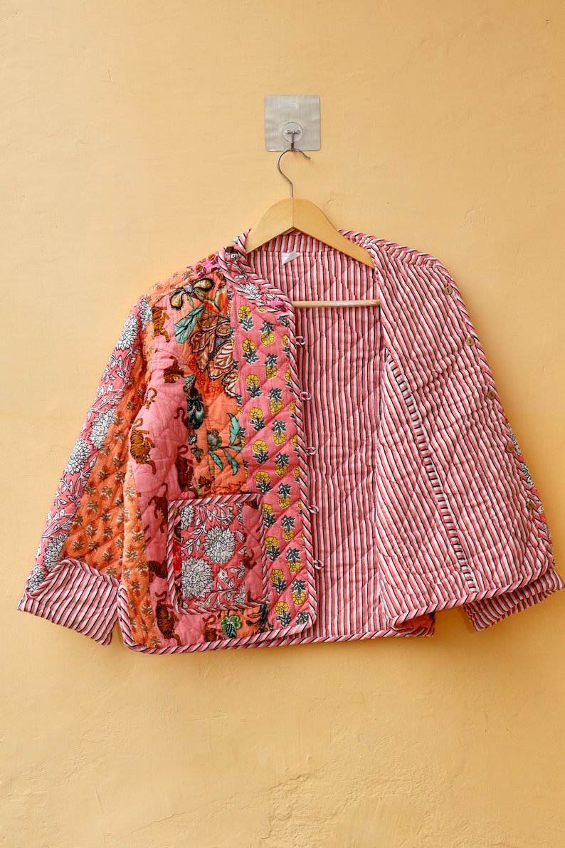 Op de afbeelding: Gewatteerde jas met een patchwork ontwerp. Het heeft bloemen- en dierenprints in roze, oranje en groen. De binnenvoering heeft rood-witte strepen. Het hangt aan een houten kleerhanger.