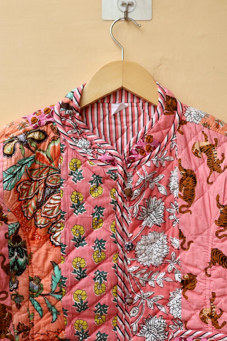 Op de afbeelding: Een gewatteerde jas met een bloemen- en tijgerprint. De jas heeft een roze achtergrond met diverse bloempatronen in wit, geel en oranje. De kraag en de bies hebben een gestreept patroon. De jas hangt aan een houten hanger.