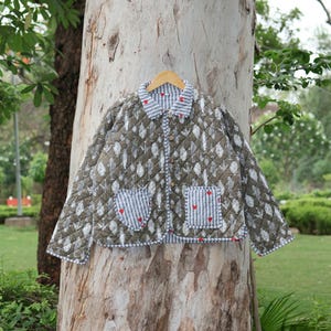 May include: A quilted jacket with a leaf pattern in shades of olive green and white, hanging on a wooden hanger. The collar and pockets feature a blue and white striped fabric with small red heart accents. The jacket is buttoned up.