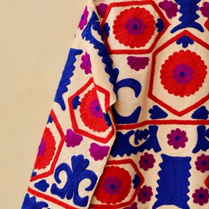 May include: A cream-colored jacket with intricate embroidery in shades of red, blue, and purple. The embroidery features geometric patterns and floral motifs.