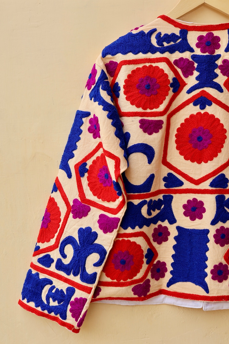 May include: A cream-colored jacket with intricate embroidery in shades of red, blue, and purple. The embroidery features geometric patterns and floral motifs.