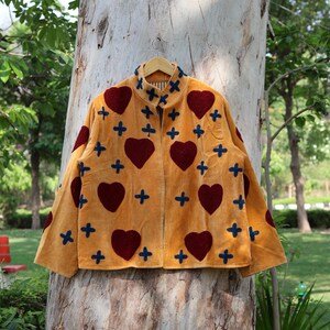 Puede incluir: Chaqueta mostaza con cuello alto, adornada con grandes corazones burdeos y cruces azules. La chaqueta est&aacute; colgada en una percha de madera, mostrando un dise&ntilde;o &uacute;nico y artesanal, con patrones de corazones y cruces.