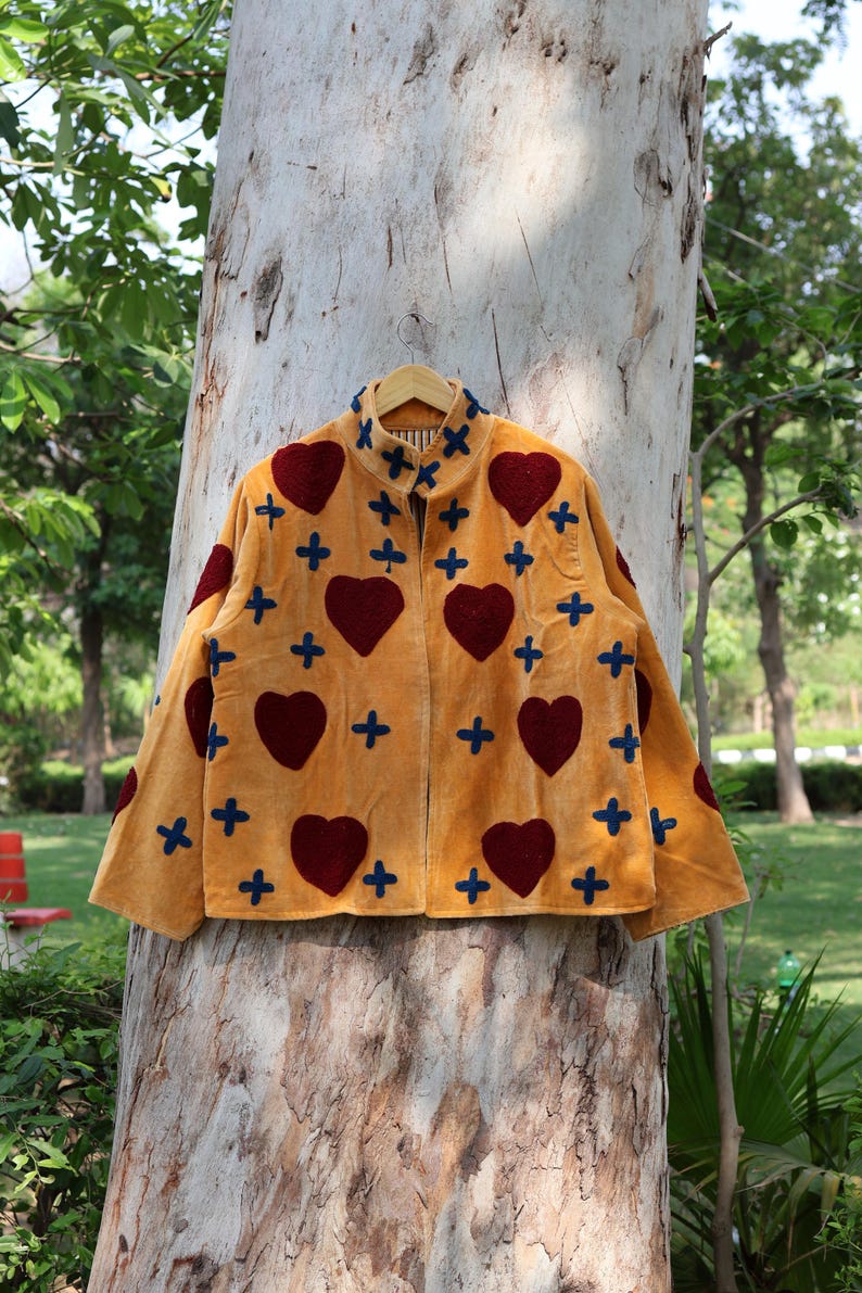Puede incluir: Chaqueta mostaza con cuello alto, adornada con grandes corazones burdeos y cruces azules. La chaqueta est&aacute; colgada en una percha de madera, mostrando un dise&ntilde;o &uacute;nico y artesanal, con patrones de corazones y cruces.