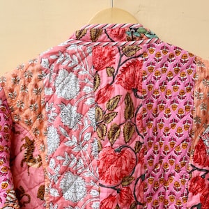 Op de afbeelding: Gewatteerde jas met een patchwork ontwerp met bloemen- en botanische patronen in roze, rode en perziktinten. De jas hangt aan een houten hanger tegen een neutrale achtergrond.