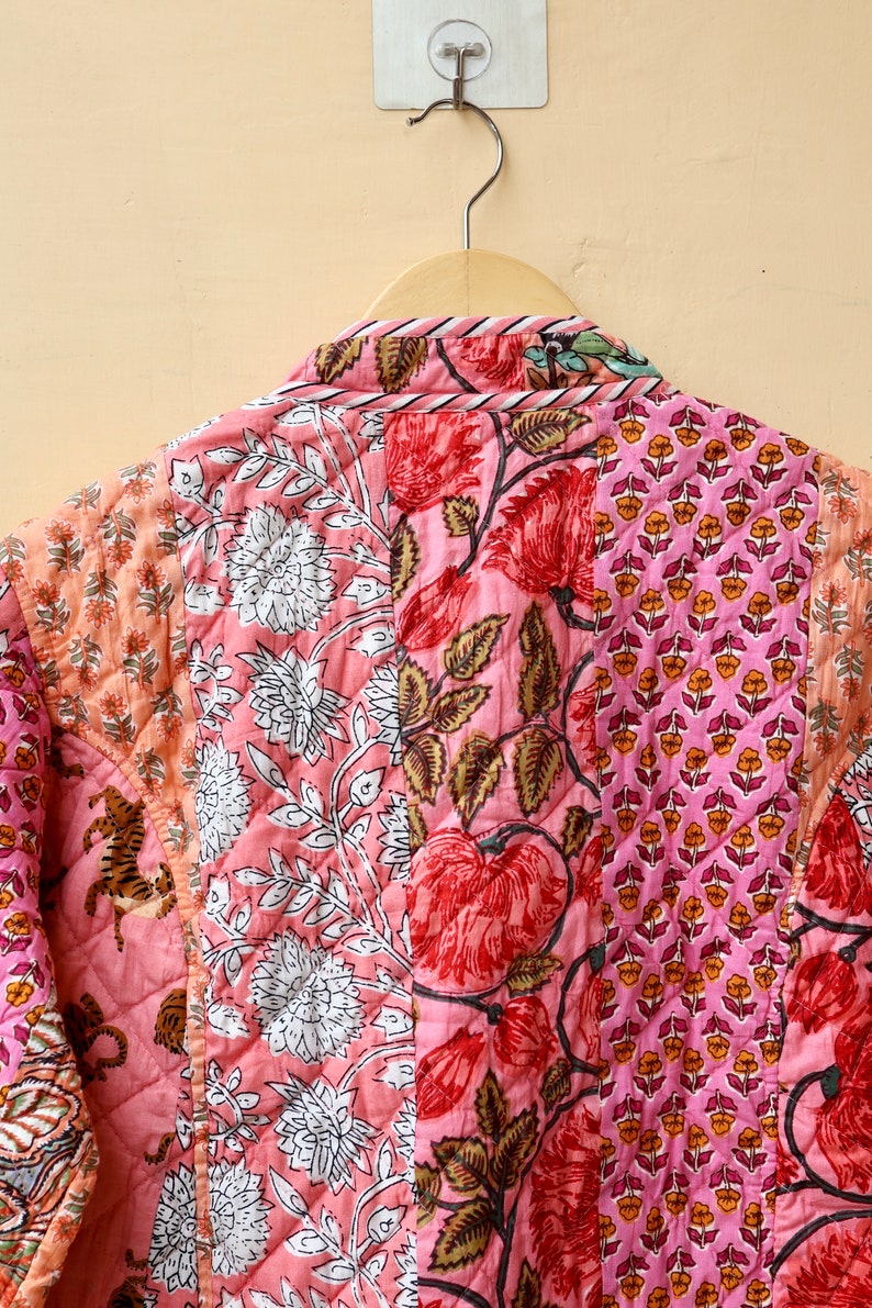 Op de afbeelding: Gewatteerde jas met een patchwork ontwerp met bloemen- en botanische patronen in roze, rode en perziktinten. De jas hangt aan een houten hanger tegen een neutrale achtergrond.