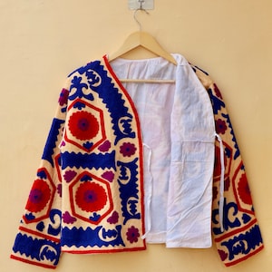 May include: A cream-colored jacket with blue, red, and purple embroidery. The jacket has a geometric pattern with floral motifs. The jacket is open and hanging on a wooden hanger.