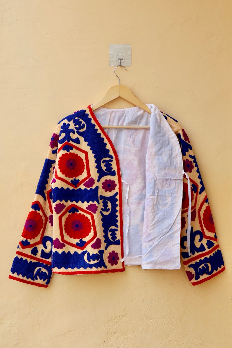May include: A cream-colored jacket with blue, red, and purple embroidery. The jacket has a geometric pattern with floral motifs. The jacket is open and hanging on a wooden hanger.