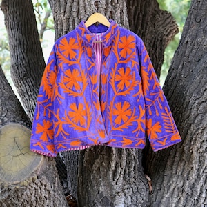 May include: A purple jacket with orange floral embroidery. The jacket has a mandarin collar and long sleeves. The interior lining is pink and white striped. The jacket is hanging on a wooden hanger against a tree trunk background.
