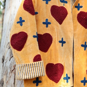 Puede incluir: Chaqueta de color amarillo mostaza con corazones rojos bordados y cruces azules. La chaqueta presenta un pu&ntilde;o a rayas y una tela suave y texturizada. Una pieza &uacute;nica, hecha a mano, para un estilo vintage o bohemio.