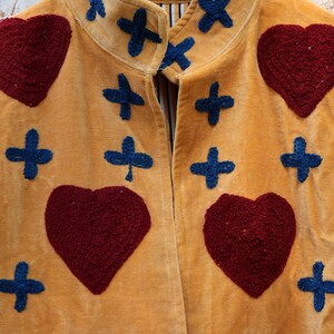 Puede incluir: Chaqueta color mostaza con textura de terciopelo, adornada con corazones burdeos bordados y formas de cruz azul marino. La chaqueta est&aacute; colgada en una percha de madera, mostrando su dise&ntilde;o y artesan&iacute;a &uacute;nicos.