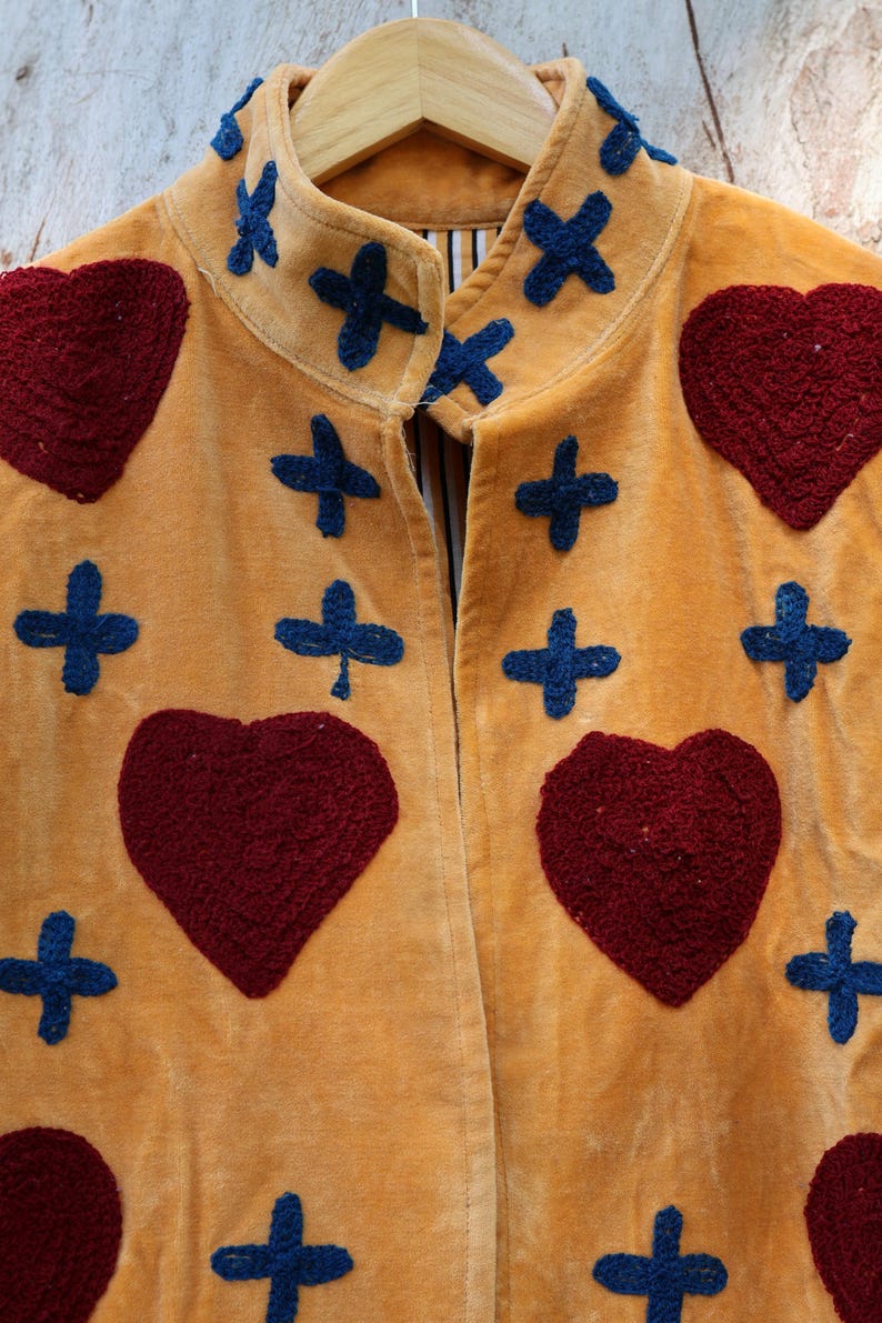 Puede incluir: Chaqueta color mostaza con textura de terciopelo, adornada con corazones burdeos bordados y formas de cruz azul marino. La chaqueta est&aacute; colgada en una percha de madera, mostrando su dise&ntilde;o y artesan&iacute;a &uacute;nicos.