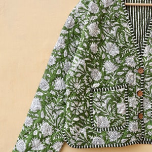 Puede incluir: Una chaqueta acolchada verde con estampado floral blanco y ribete negro. La chaqueta tiene cierre de botones y un bolsillo en el lado izquierdo.