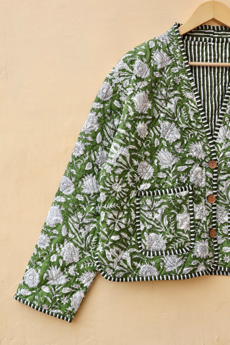 Puede incluir: Una chaqueta acolchada verde con estampado floral blanco y ribete negro. La chaqueta tiene cierre de botones y un bolsillo en el lado izquierdo.