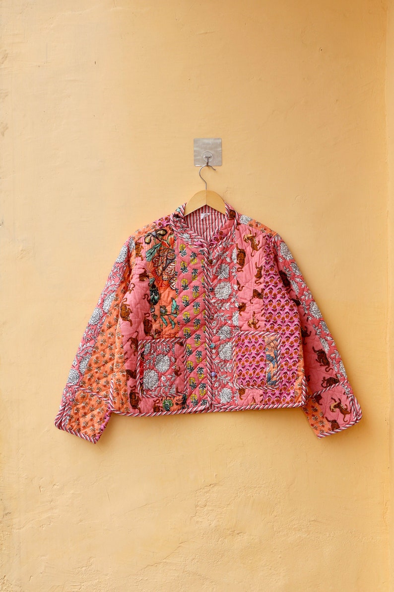 Op de afbeelding: Een gewatteerde jas in roze en oranje tinten, met een patchwork ontwerp met bloemen- en diermotieven. De jas heeft een gestreepte kraag en bies, en hangt aan een houten hanger.