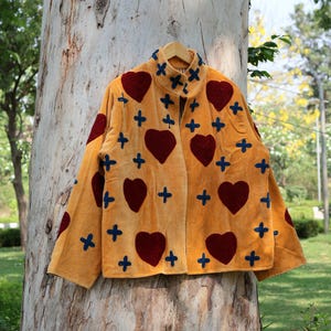 Puede incluir: Chaqueta color mostaza con cuello alto, adornada con grandes corazones burdeos y detalles de cruces azul marino. La chaqueta est&aacute; colgada en una percha de madera contra un tronco de &aacute;rbol, mostrando un dise&ntilde;o &uacute;nico y artesanal.